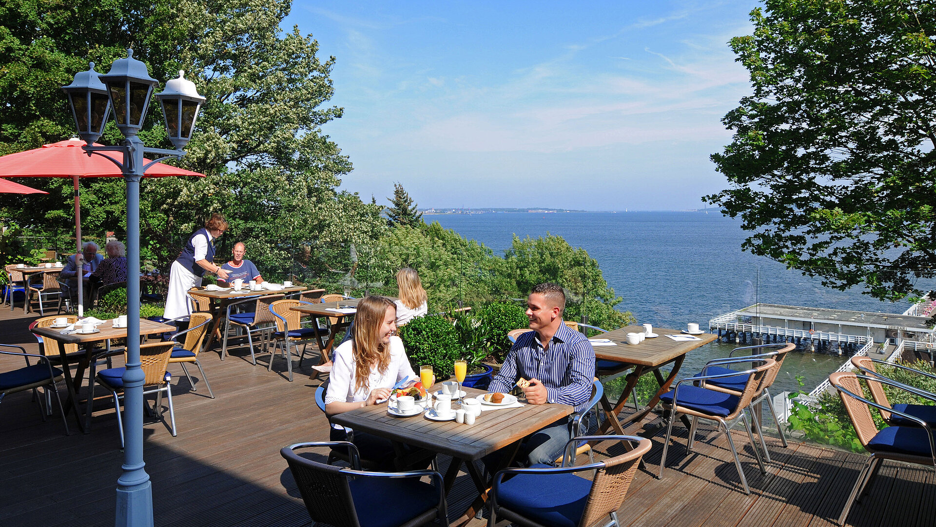 Panorama terrace at the Maritim Hotel Kiel with guests at breakfast, sunny weather and view of the fjord in the background.