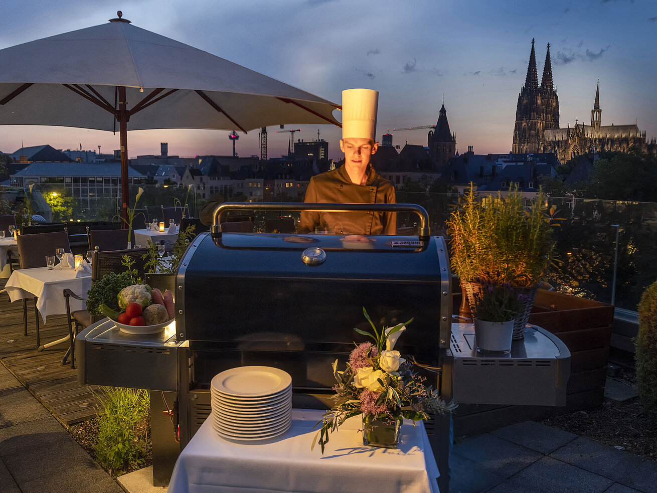 A chef grilling in the evening on the rooftop terrace with the illuminated Cologne Cathedral in the background.