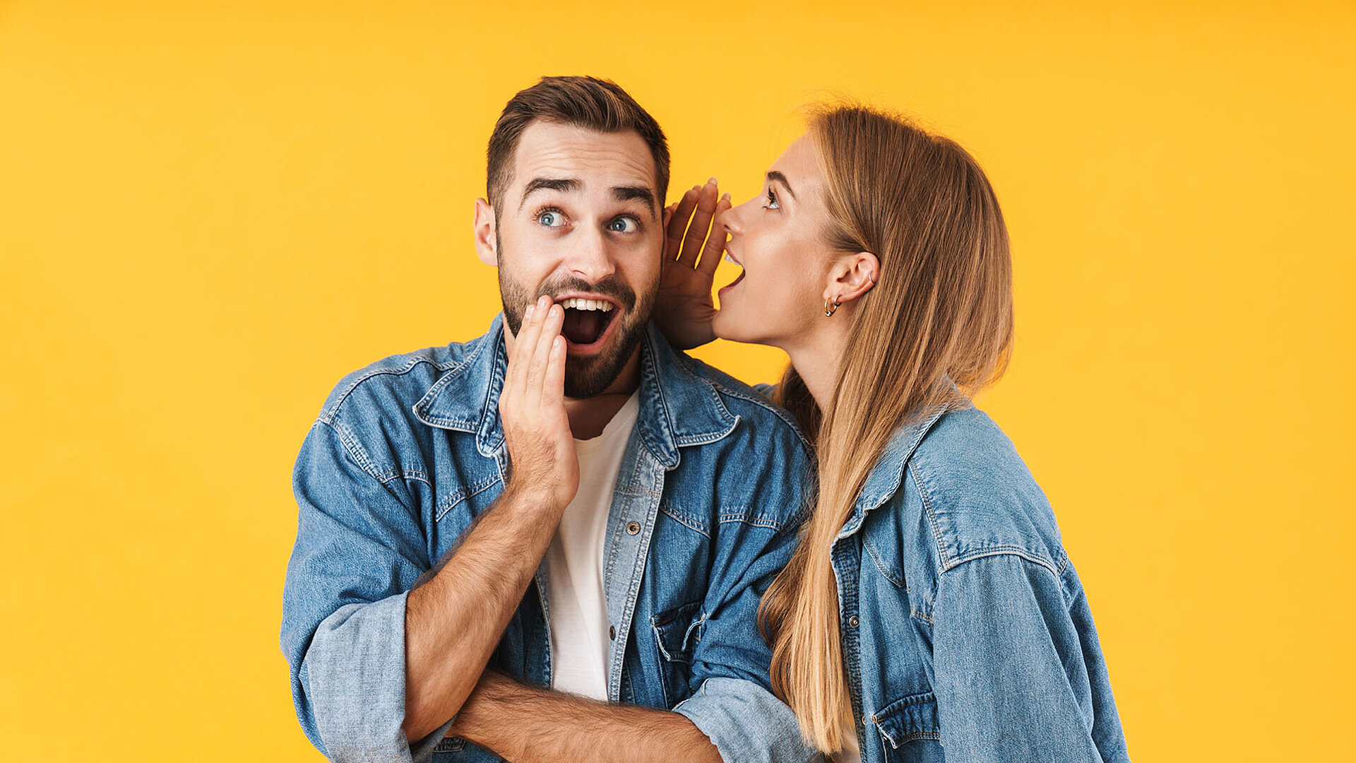 Woman whispers secret to man, both laughing in denim jackets against yellow background