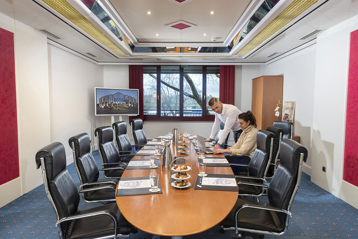 Dos personas trabajan en el Salon Boardroom del Maritim Colonia con portátiles en una mesa de conferencia con bebidas y snacks