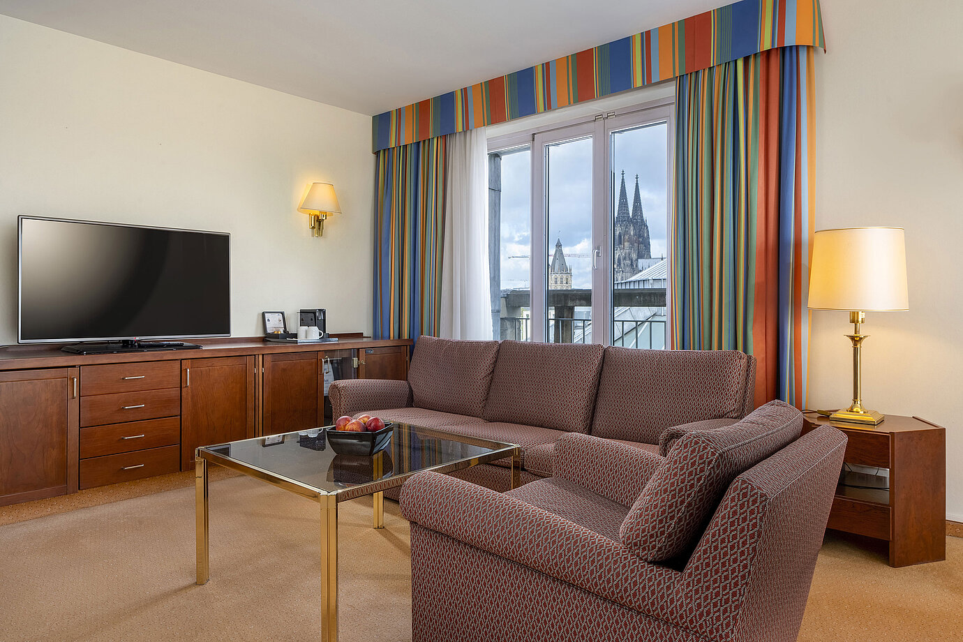 Living room of Senior Suite at Maritim Hotel Köln with sofa, TV, and Cologne Cathedral view through the windows.