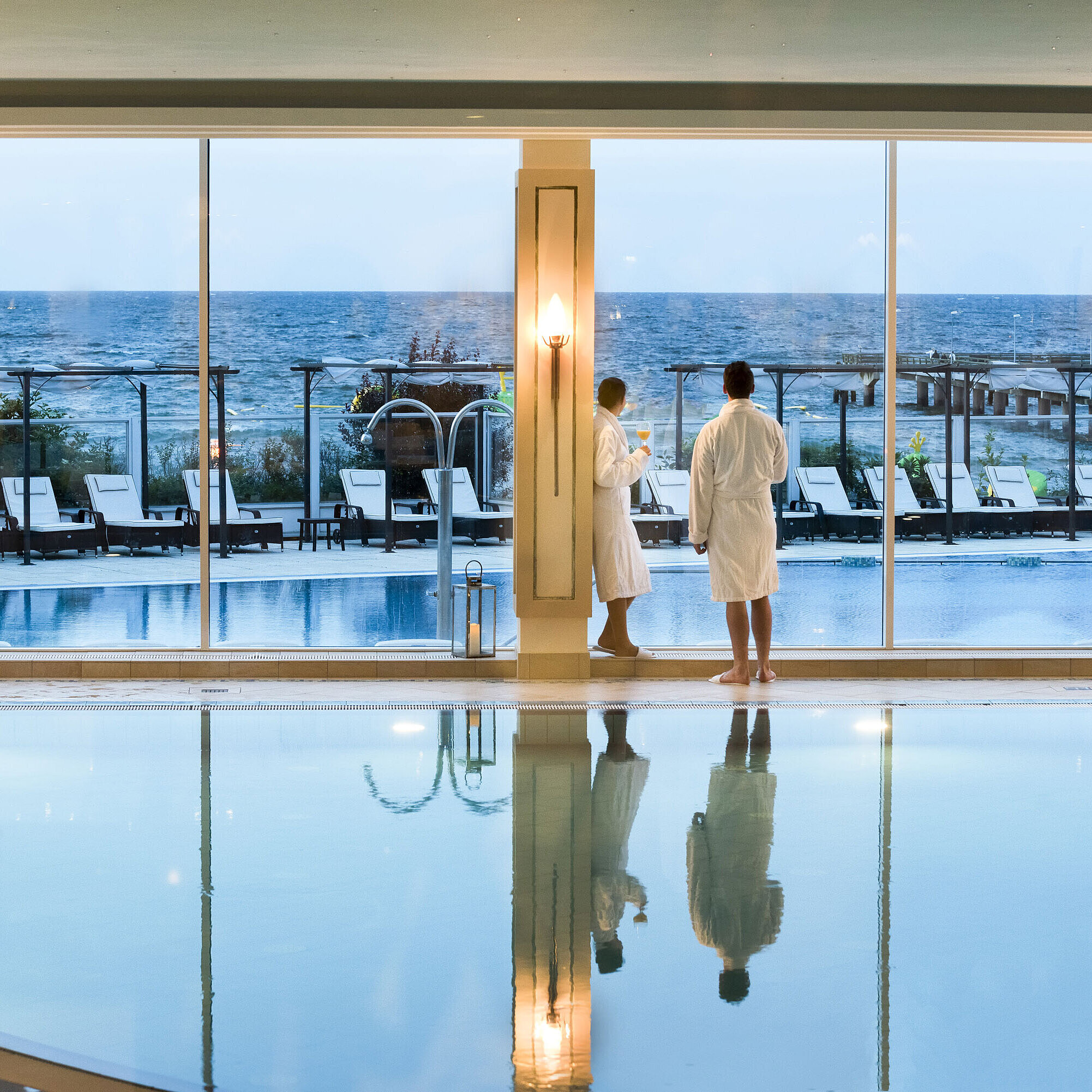 Piscina interna con vista mare al Maritim Seehotel Timmendorfer Strand, due ospiti in accappatoi alla finestra.