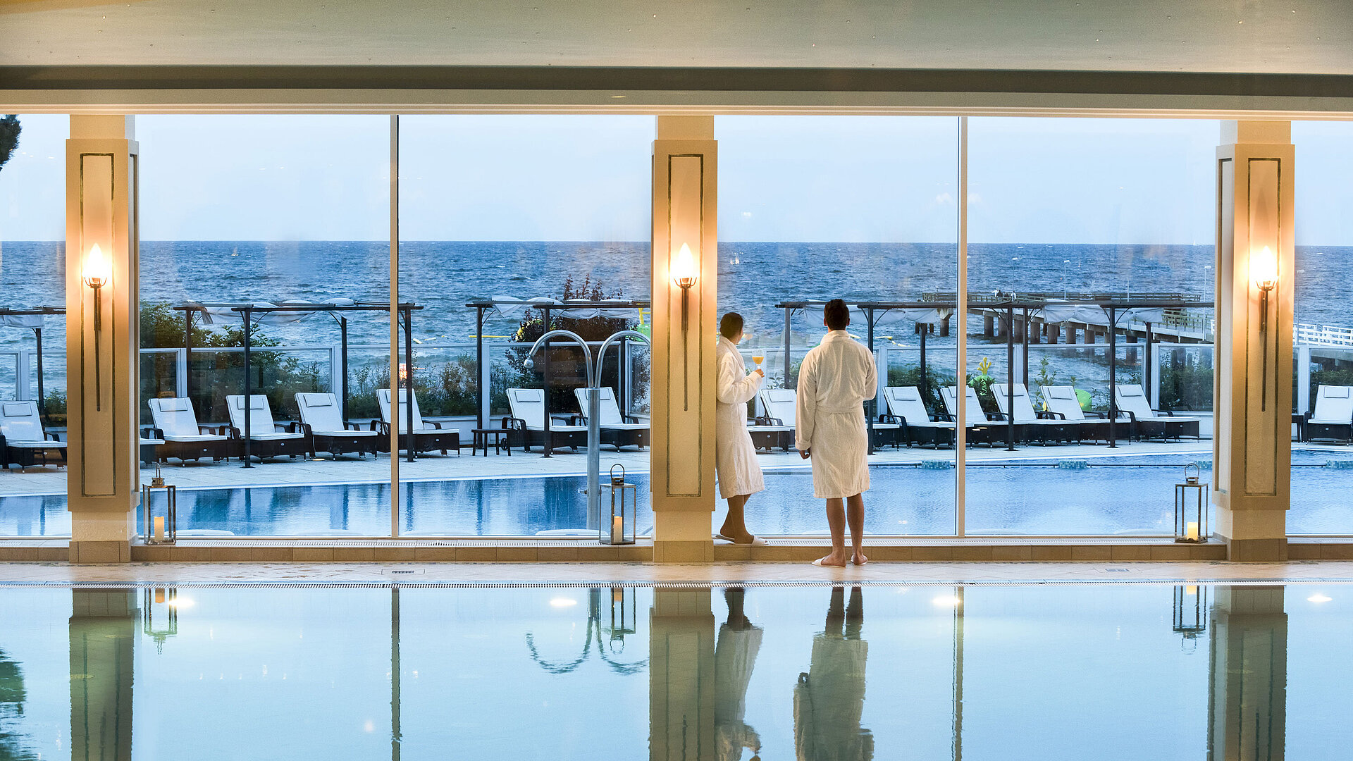 Piscina interna con vista mare al Maritim Seehotel Timmendorfer Strand, due ospiti in accappatoi alla finestra.