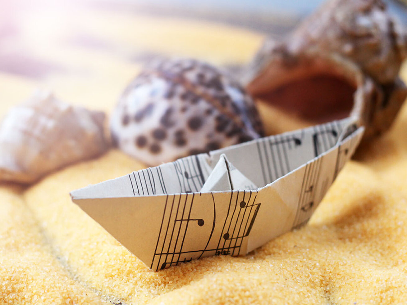 © sabdiz - AdobeStock.com Folded paper boat on decorative sand with seashells and maritime table décor.
