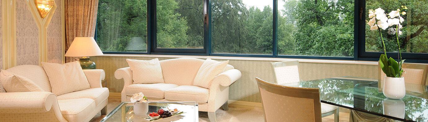 Luxurious Suite with elegant seating, glass dining table, and large panoramic window with greenery view.