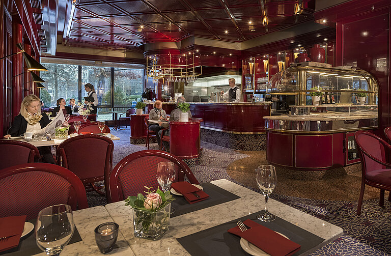 La imagen muestra el acogedor café "Brasserie" en el Hotel Maritim Bonn con decoración roja y un ambiente relajado.