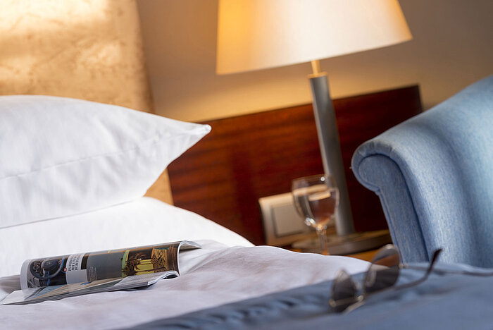 Primo piano di una camera al Maritim Hotel Fulda con letto, rivista, occhiali e bicchiere d’acqua sul comodino.