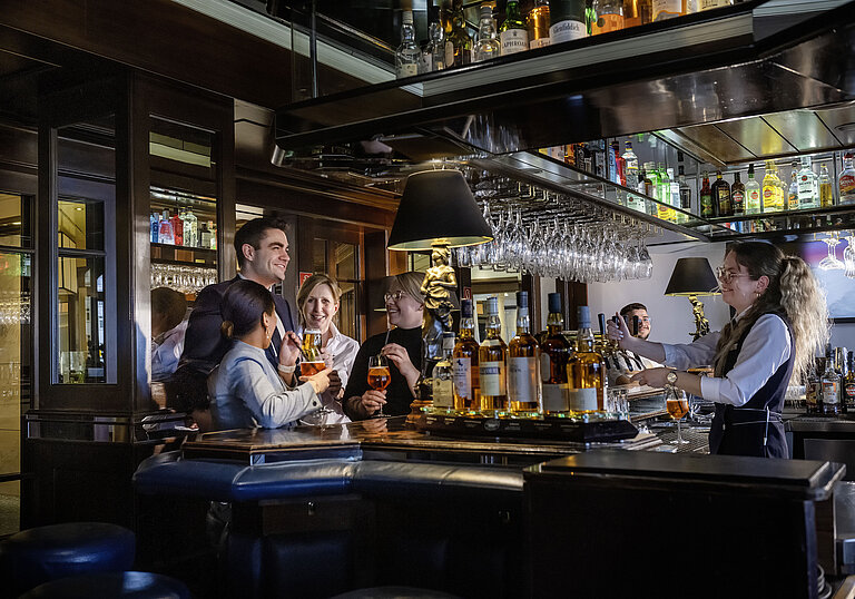 Huéspedes disfrutando de cócteles y buen ambiente en el bar del Hotel Maritim Darmstadt. Personal amable sirviendo bebidas.