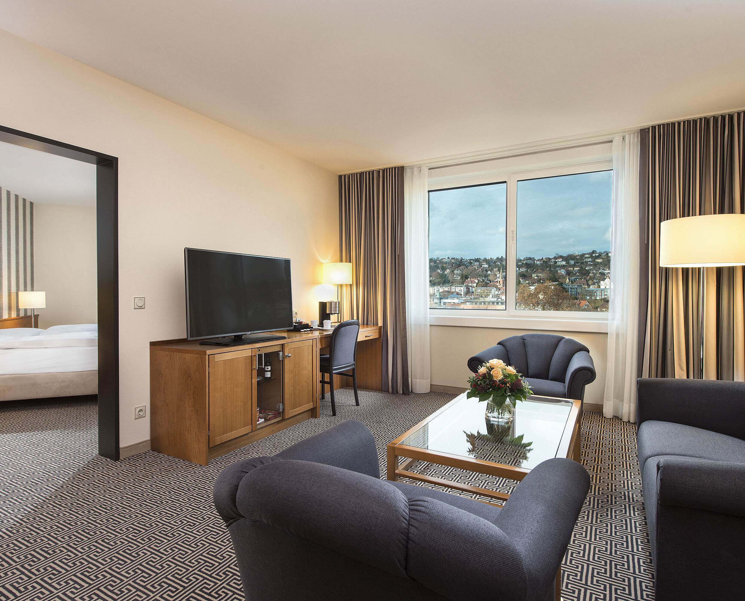 Bright suite with city view, sofas, TV, desk and bedroom at the Maritim Hotel Stuttgart