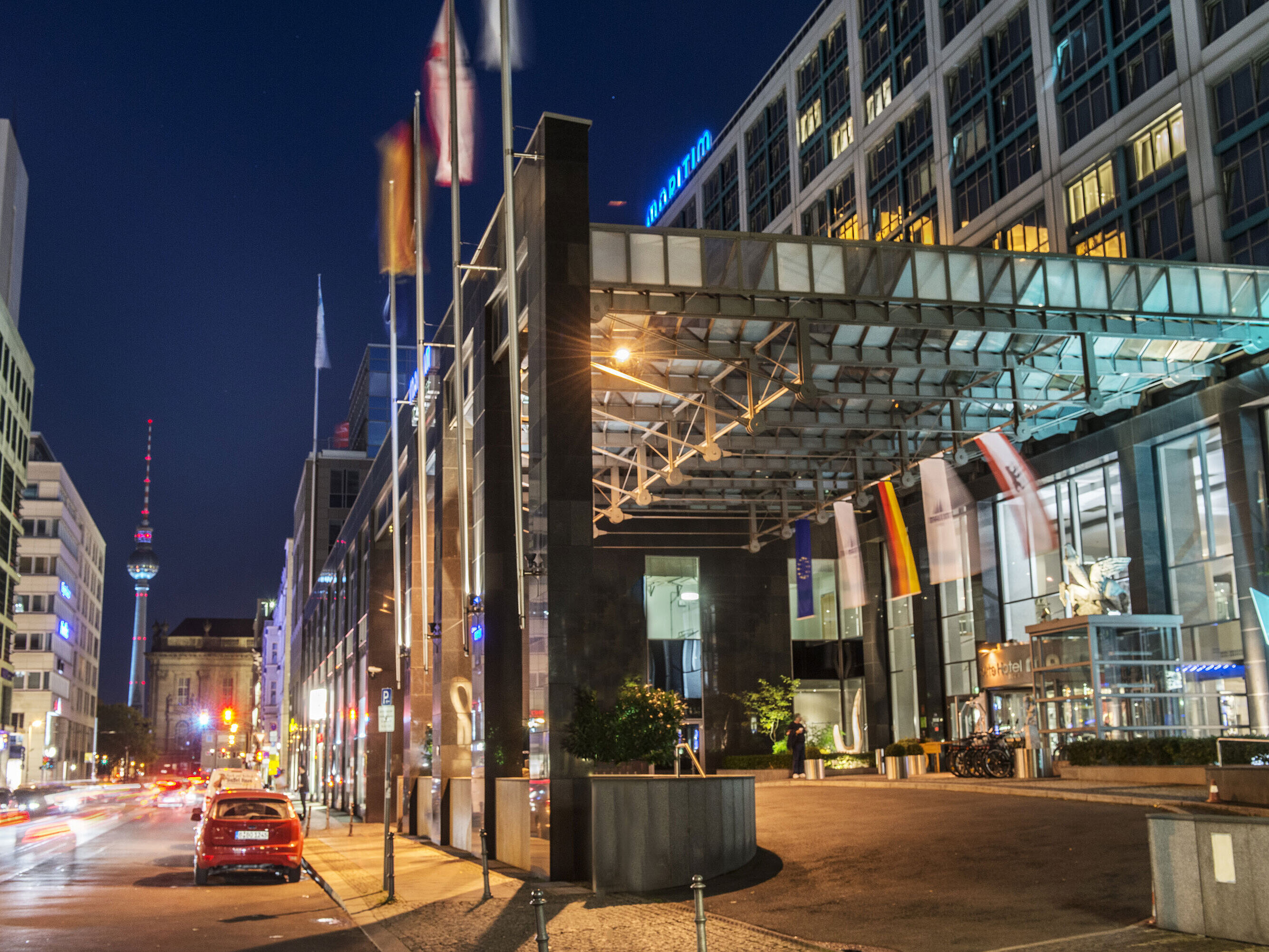 Vista notturna dell'entrata illuminata del Maritim proArte Hotel Berlino con torre della TV sullo sfondo e bandiere.