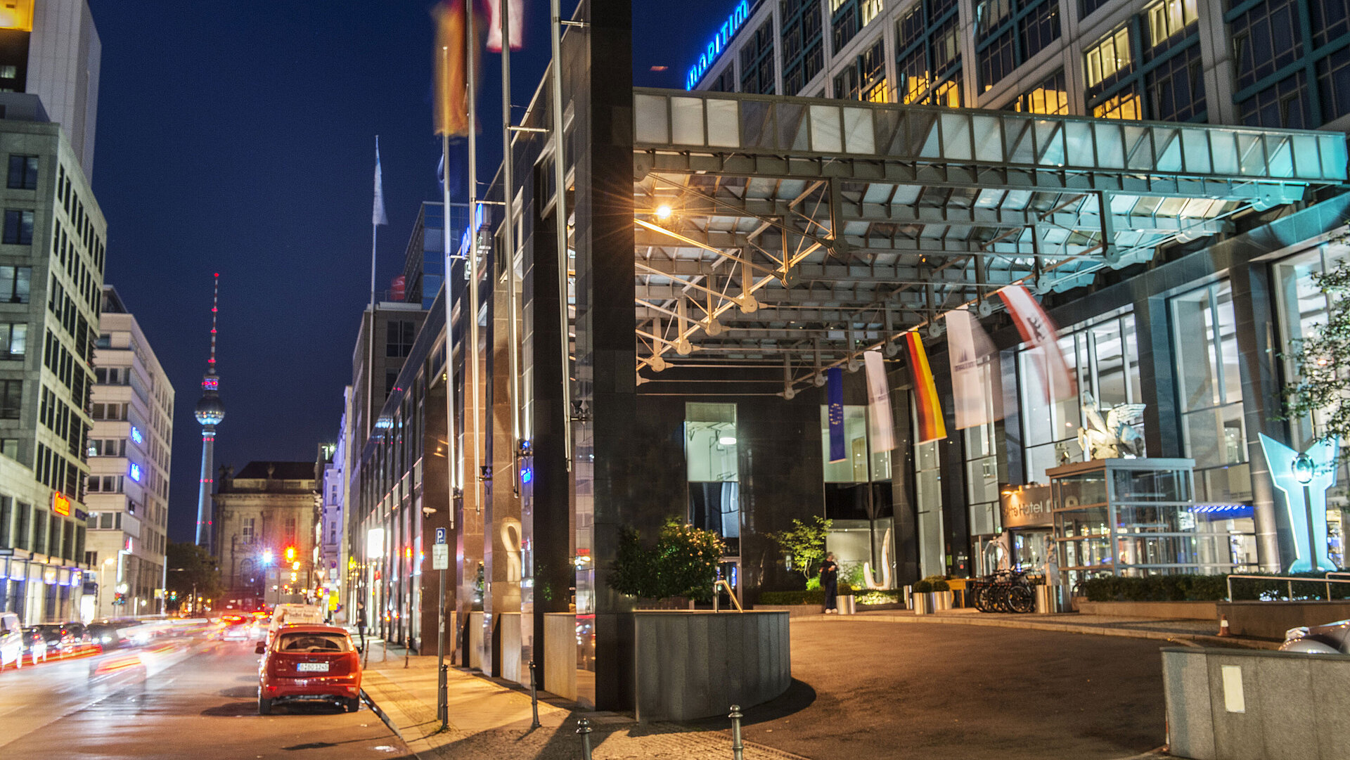 Vista notturna dell'entrata illuminata del Maritim proArte Hotel Berlino con torre della TV sullo sfondo e bandiere.