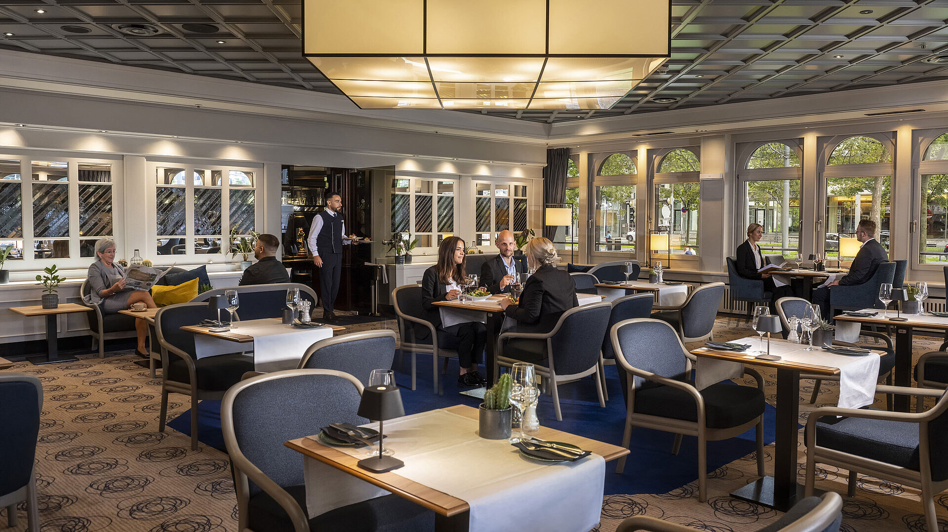 Elegant restaurant at Maritim Hotel Darmstadt with guests dining and relaxing. Modern decor and large windows with a view.