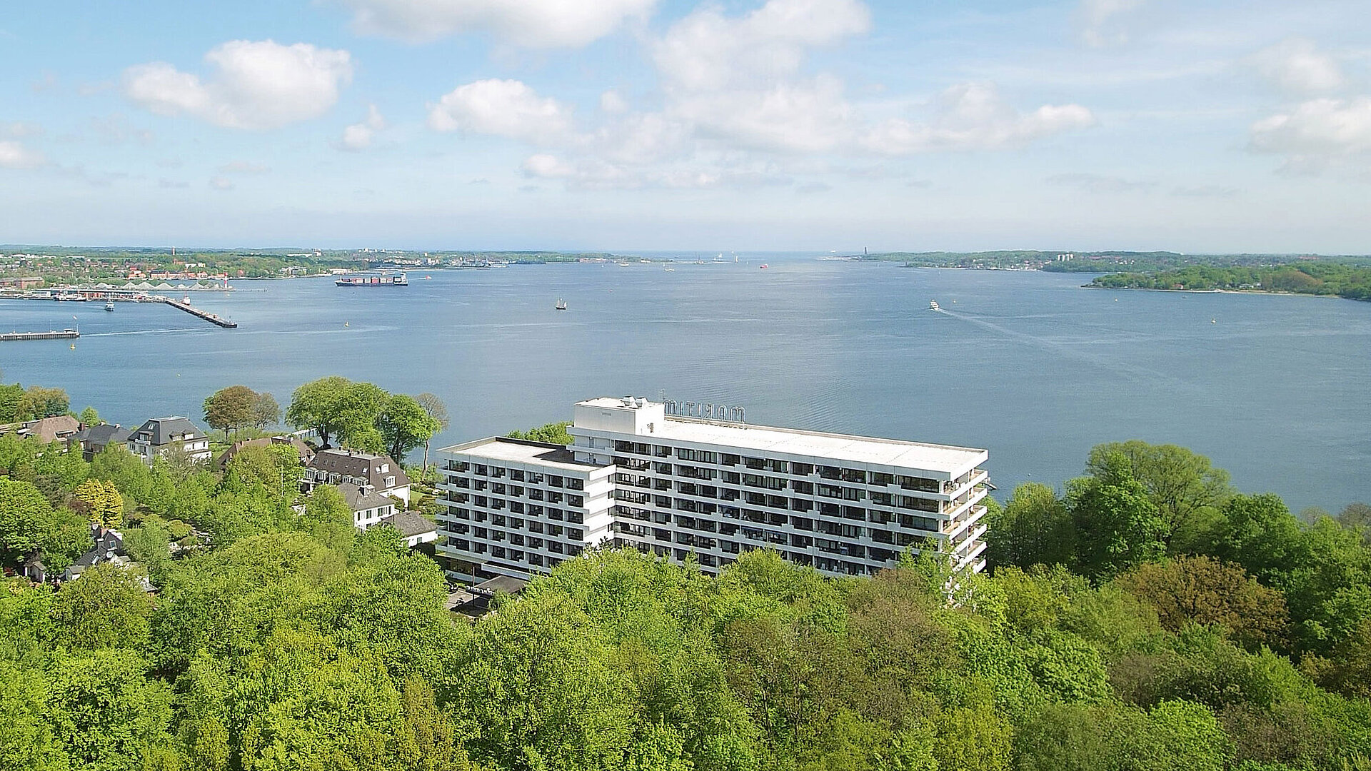 Aerial view of the Maritim Hotel Kiel, surrounded by green countryside and the Kiel Fjord, under a bright blue sky.