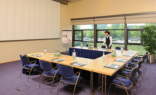 Salle de réunion de l'hôtel Maritim de Dresde avec tables en U, écran de présentation et préparations pour une réunion.