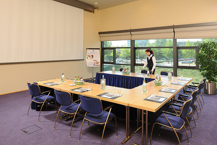 Sala de reuniones del Maritim Hotel Dresde con mesas en U, pantalla de presentación y preparativos para una reunión.