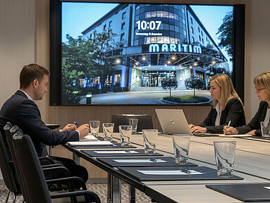 Meeting room with conference table, participants in discussion and a presentation shown on the screen.