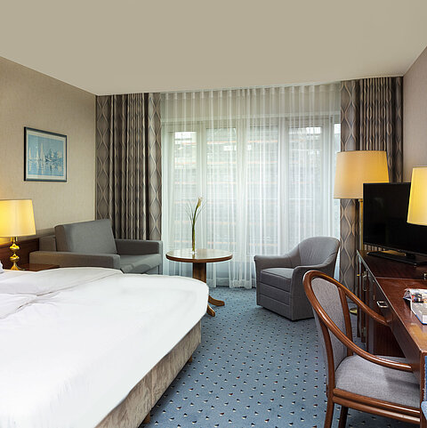 Bright family room at Maritim Hotel Cologne with double bed, desk, TV, armchairs, and large windows with curtains.