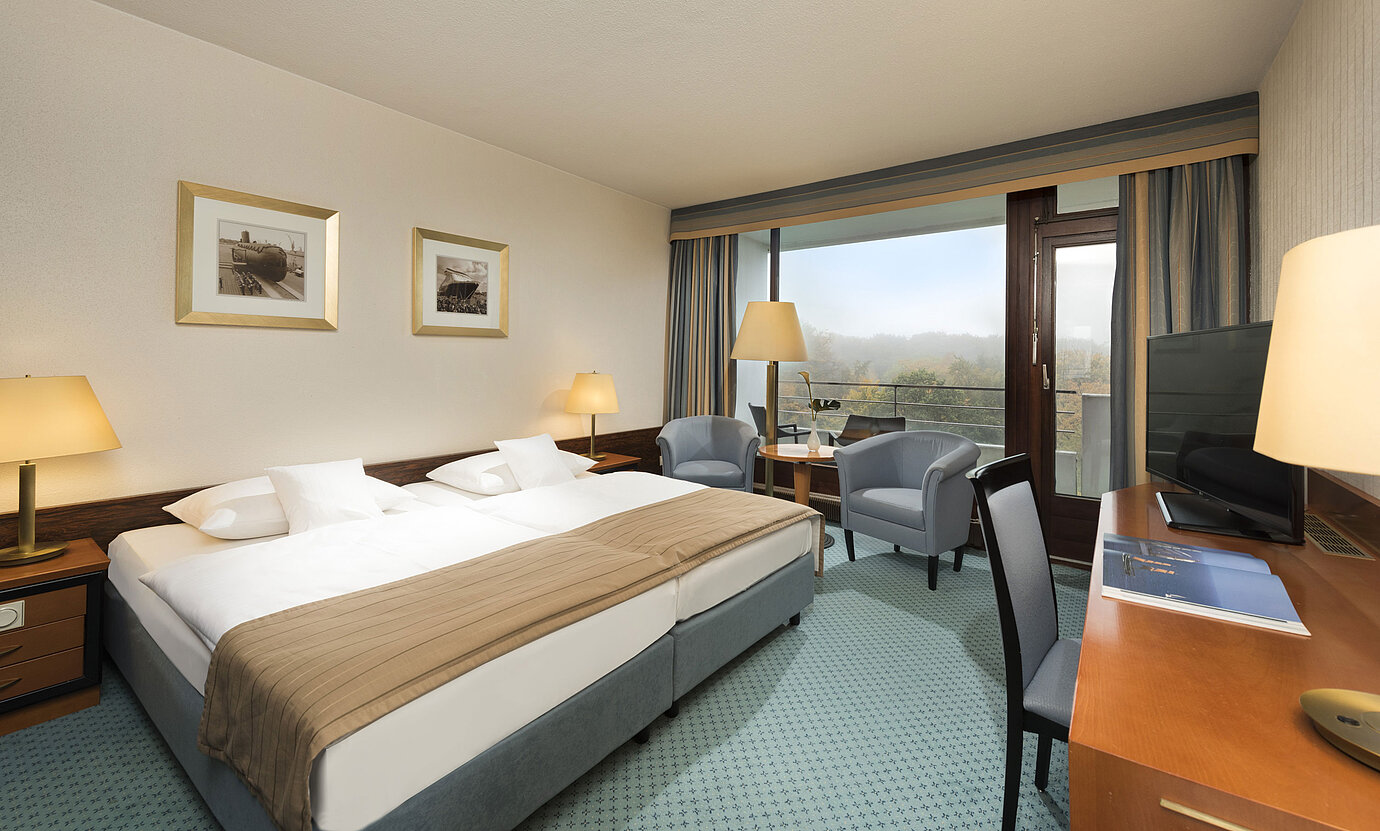 Chambre Classic du Maritim Hotel Kiel avec grand lit double, coin salon, bureau et balcon avec vue sur la verdure.