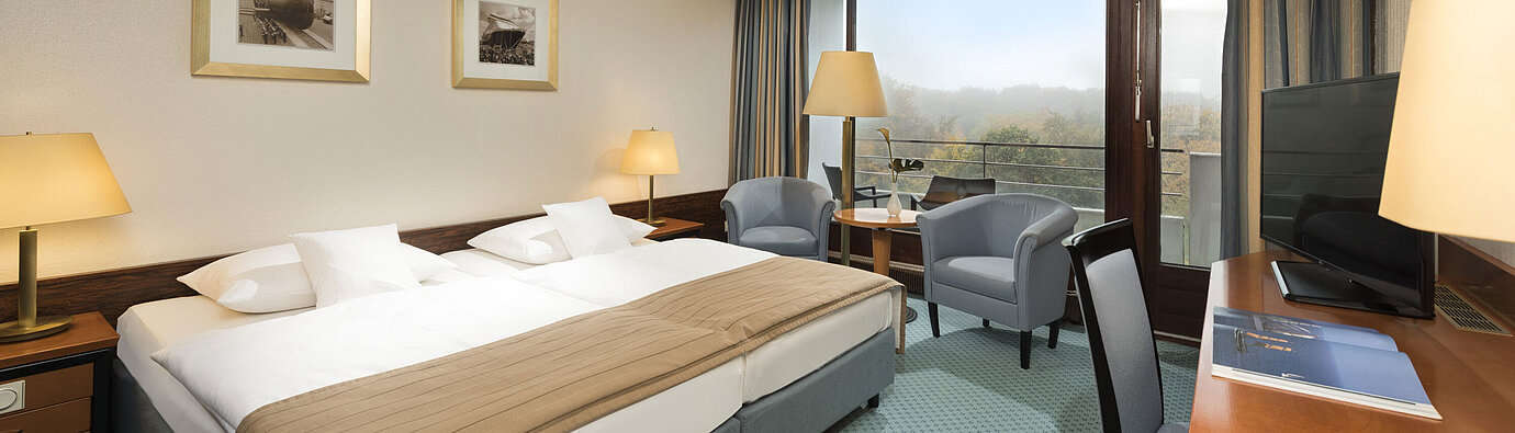 Camera classica del Maritim Hotel Kiel con ampio letto matrimoniale, area salotto, scrivania e balcone con vista sul verde.