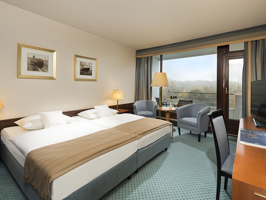 Camera classica del Maritim Hotel Kiel con ampio letto matrimoniale, area salotto, scrivania e balcone con vista sul verde.