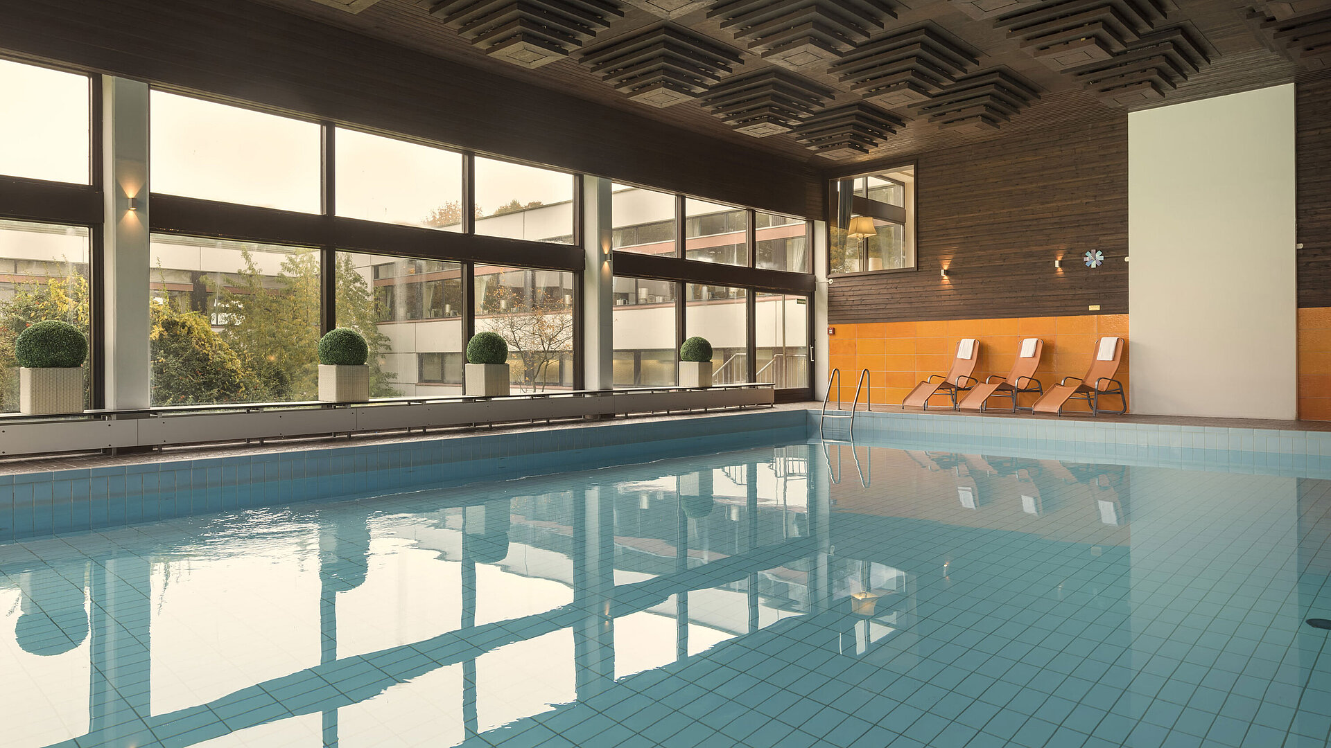 La photo montre la piscine intérieure du Maritim Hotel Kiel, entourée de grandes fenêtres, avec des chaises longues pour la détente.