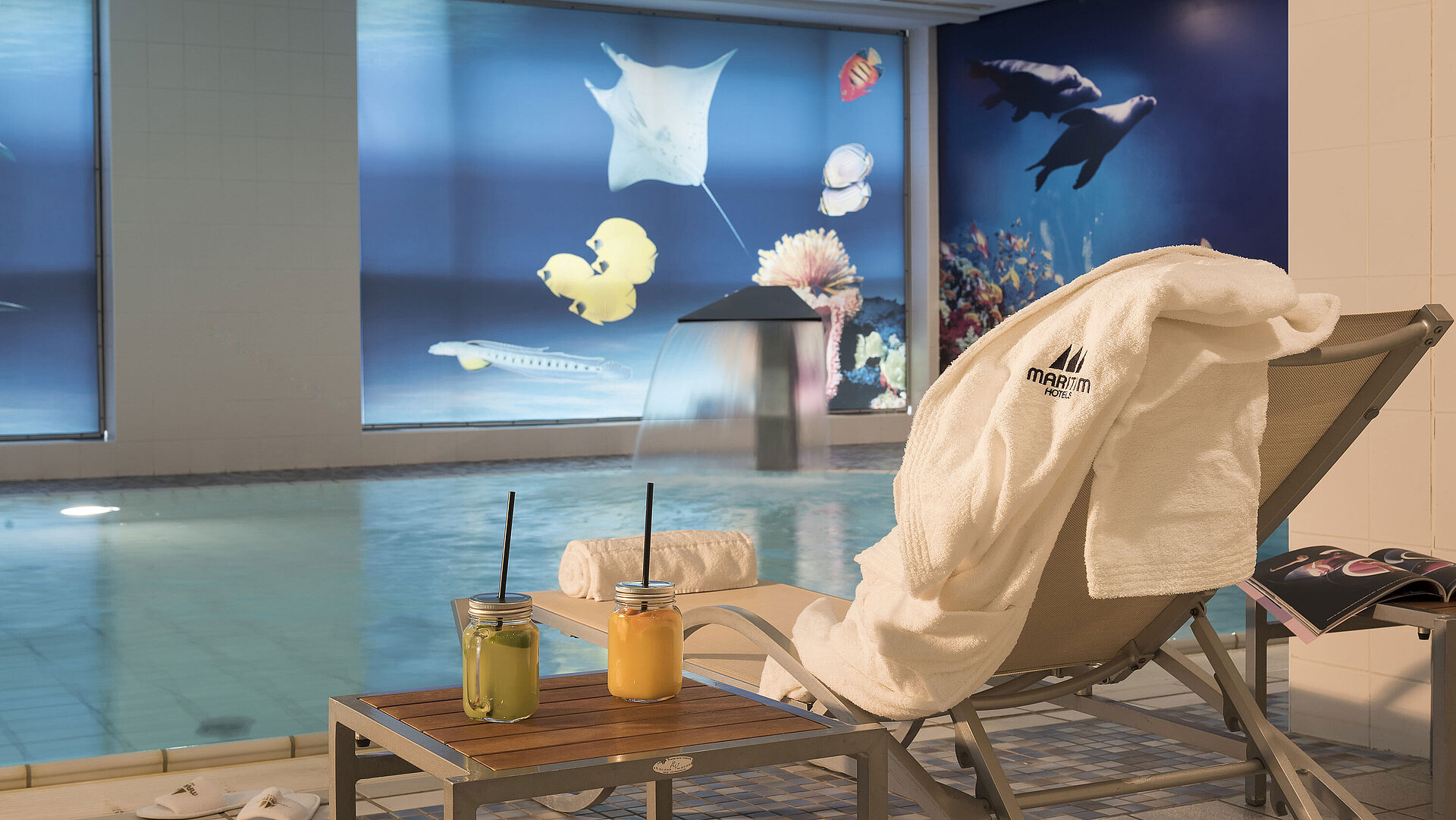 Area relax vicino alla piscina del Maritim proArte Hotel Berlino con sdraio,asciugamano e succhi freschi accanto a una cascata