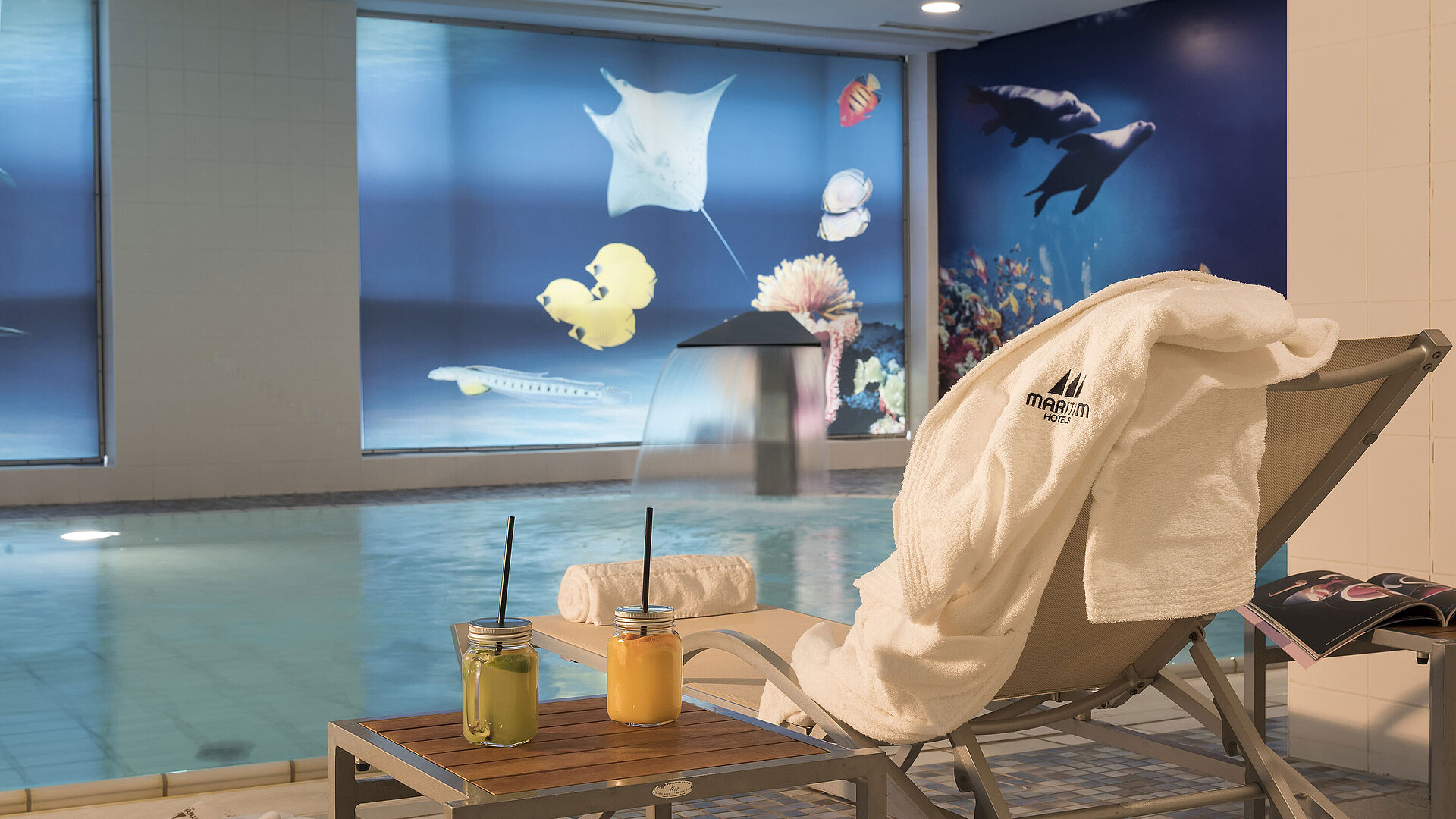 Area relax vicino alla piscina del Maritim proArte Hotel Berlino con sdraio,asciugamano e succhi freschi accanto a una cascata