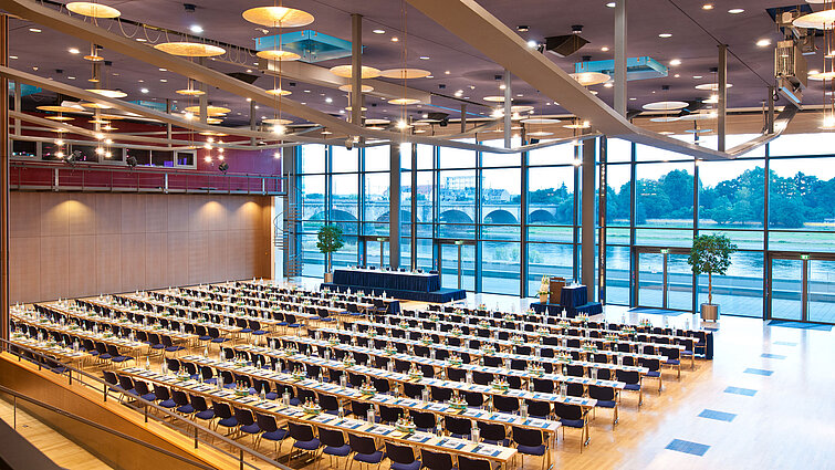 Grande salle de conférence de l'hôtel Maritim de Dresde avec des rangées de chaises bleues et vue sur l'Elbe.