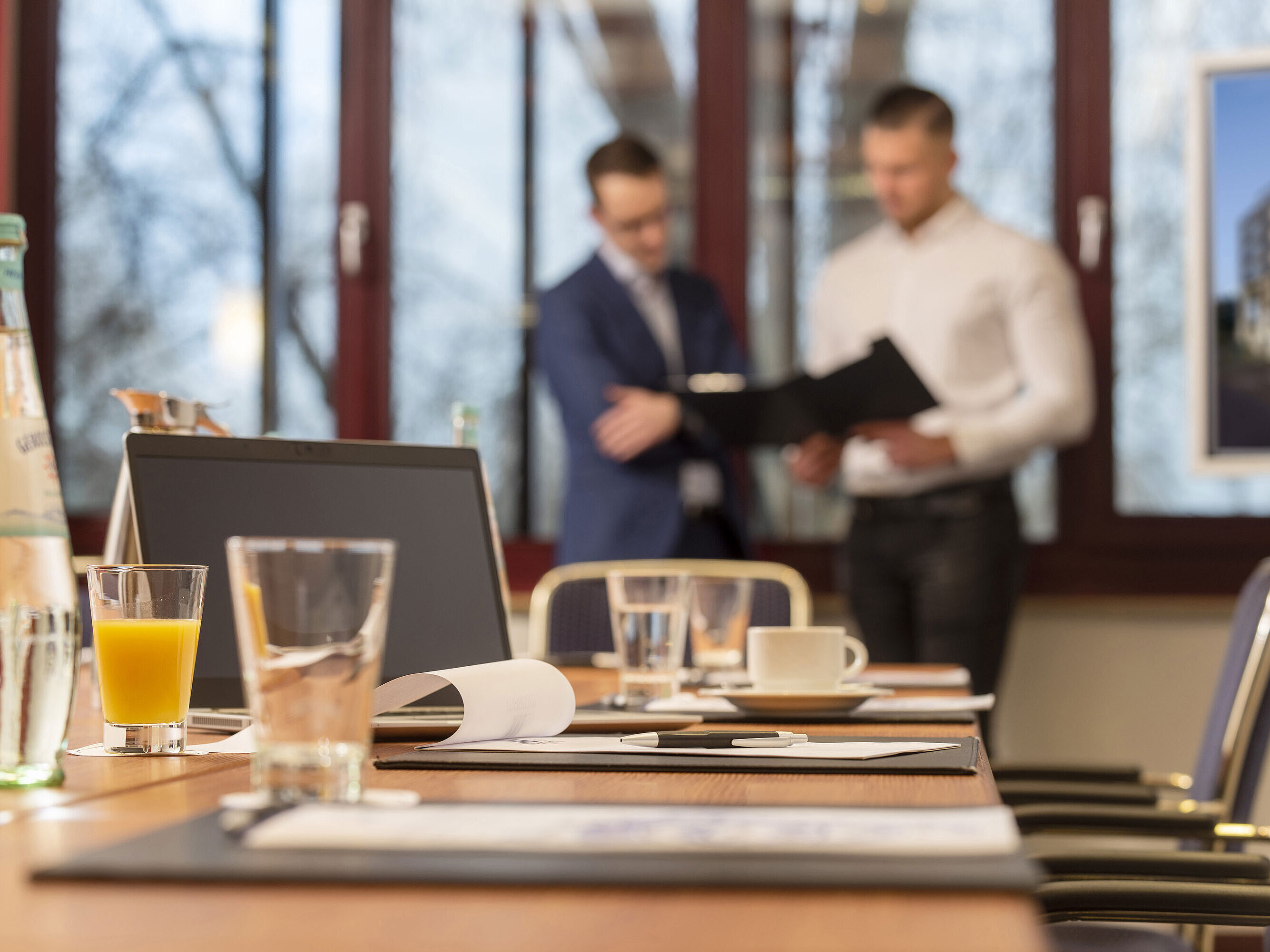 Conference room with a laptop, drinks, and documents, two businessmen in the background discussing papers.