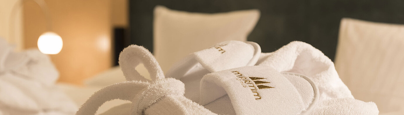 Accappatoio Accappatoio bianco e pantofole con logo Maritim sul letto, ideali per momenti di relax nella camera d’hotel