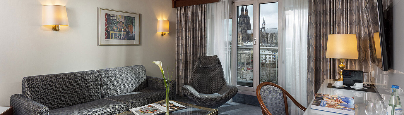Junior Suite at Maritim Hotel Cologne with a view of Cologne Cathedral, stylish sofa, chair, and desk with reading material.