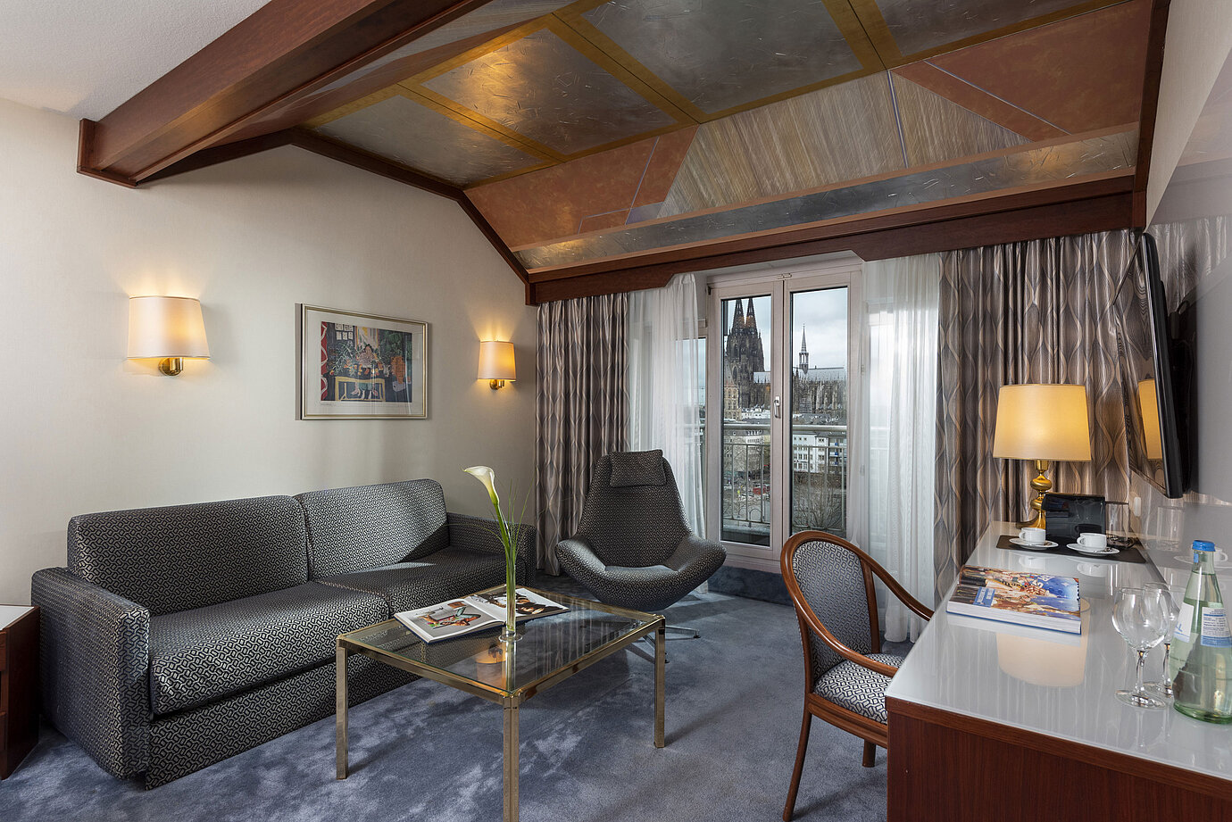 Junior Suite at Maritim Hotel Cologne with a view of Cologne Cathedral, stylish sofa, chair, and desk with reading material.