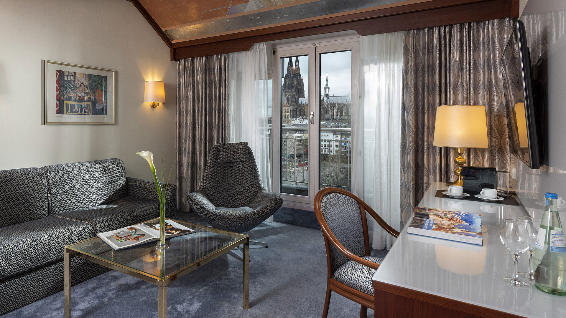 Junior Suite at Maritim Hotel Cologne with a view of Cologne Cathedral, stylish sofa, chair, and desk with reading material.
