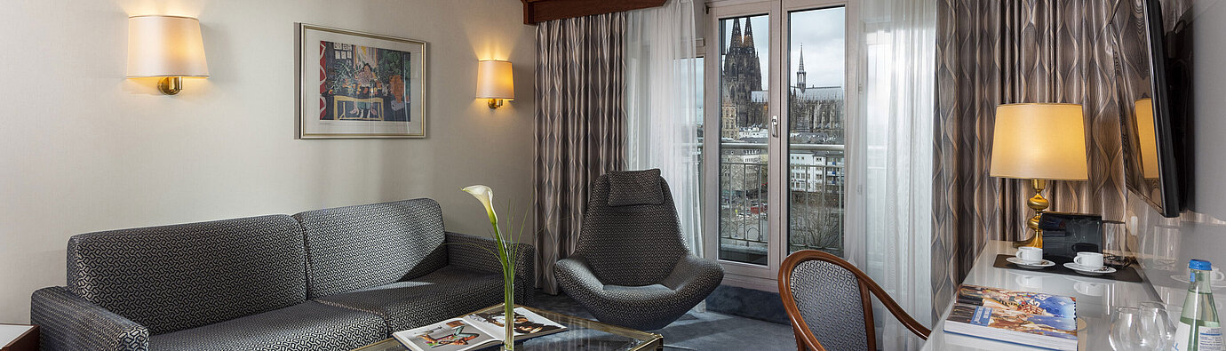 Junior Suite at Maritim Hotel Cologne with a view of Cologne Cathedral, stylish sofa, chair, and desk with reading material.