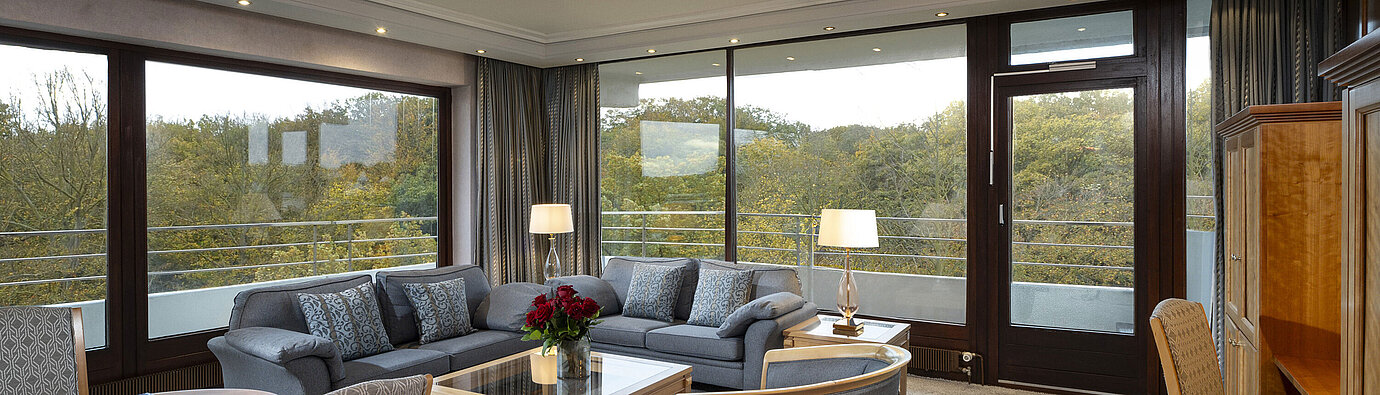 Salon de la Grand Suite de l'hôtel Maritim Kiel avec grandes fenêtres, décoration élégante et vue sur la verdure.